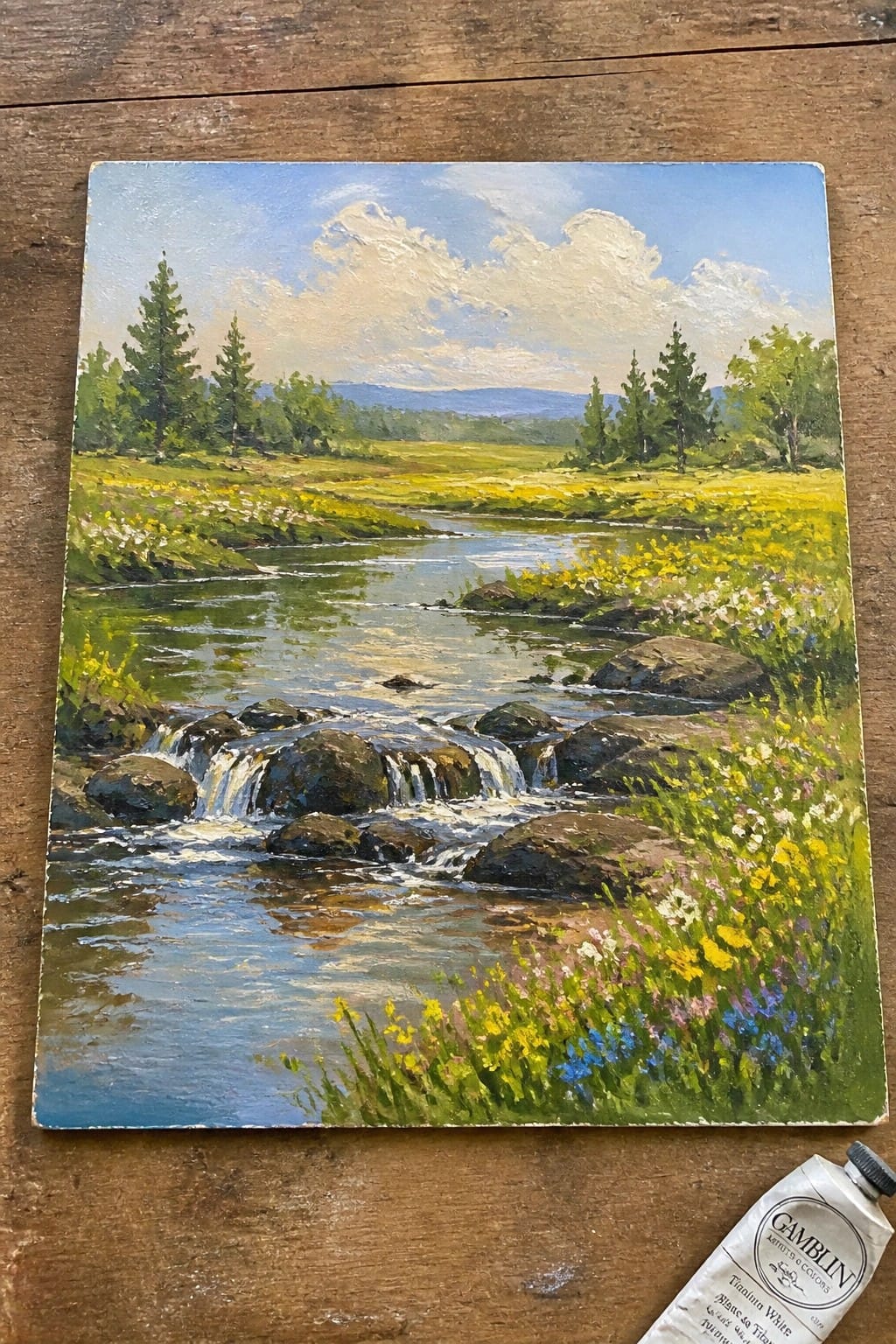 Oil painting of rocky stream through wildflower meadow beside white paint tube on wood