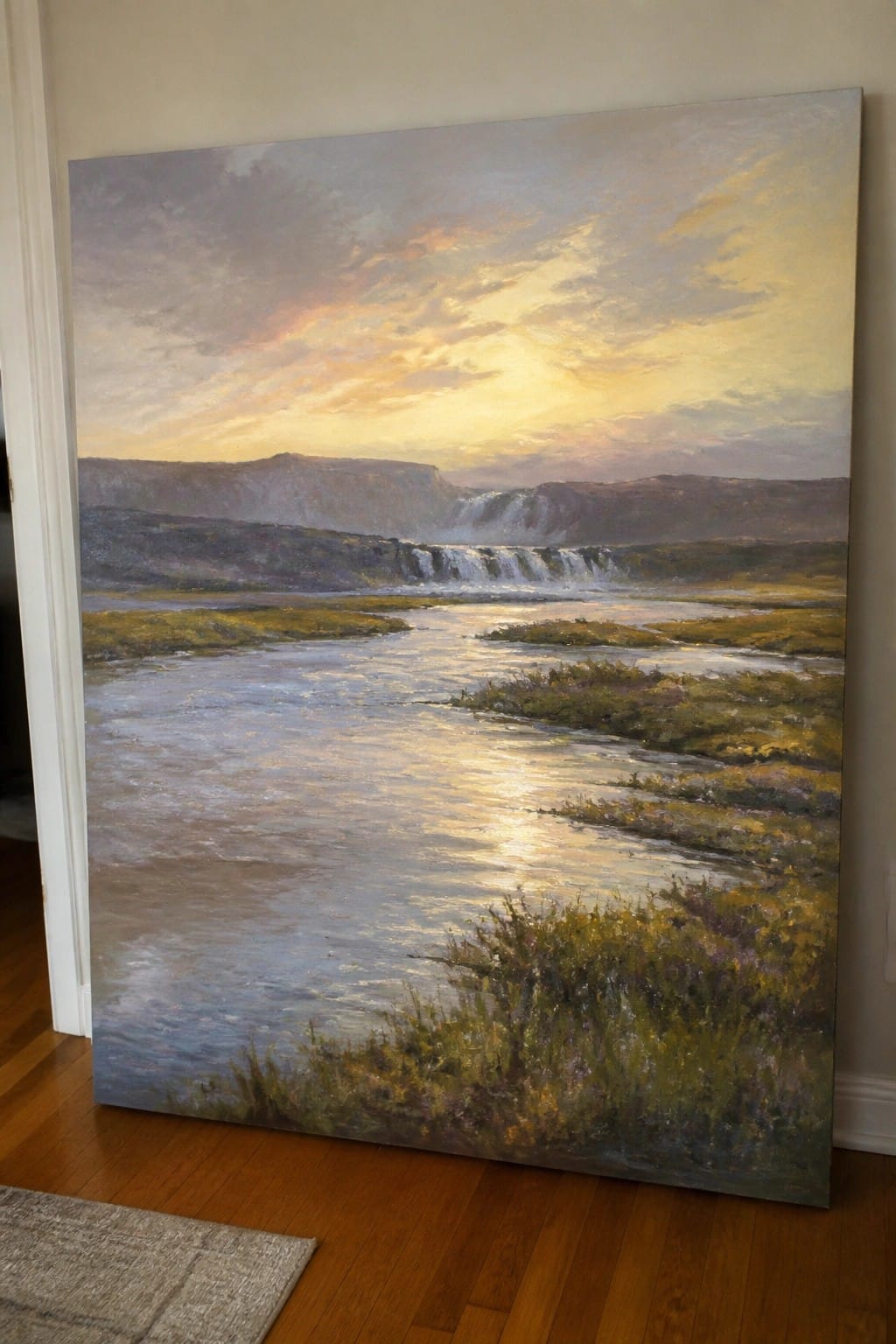 Large landscape painting of river and waterfall at sunset leaning on wall indoors.
