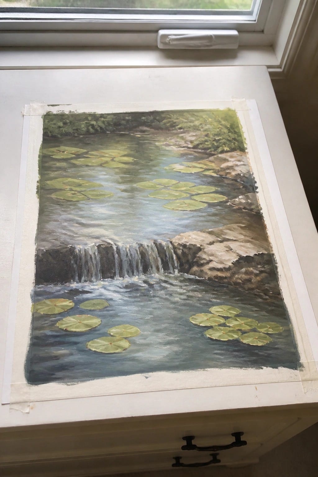 Watercolor painting of waterfall with rocks and green lily pads taped on white windowsill
