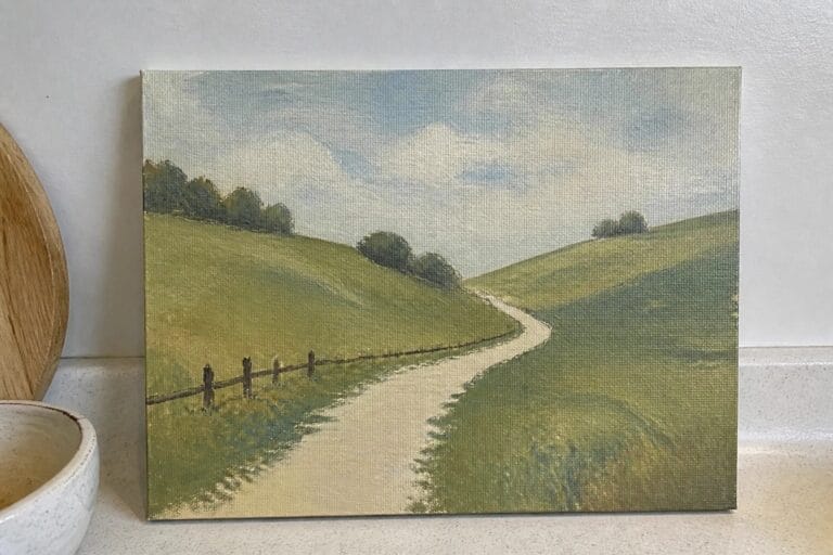Landscape painting of winding dirt road through green hills beside wooden board and jar.