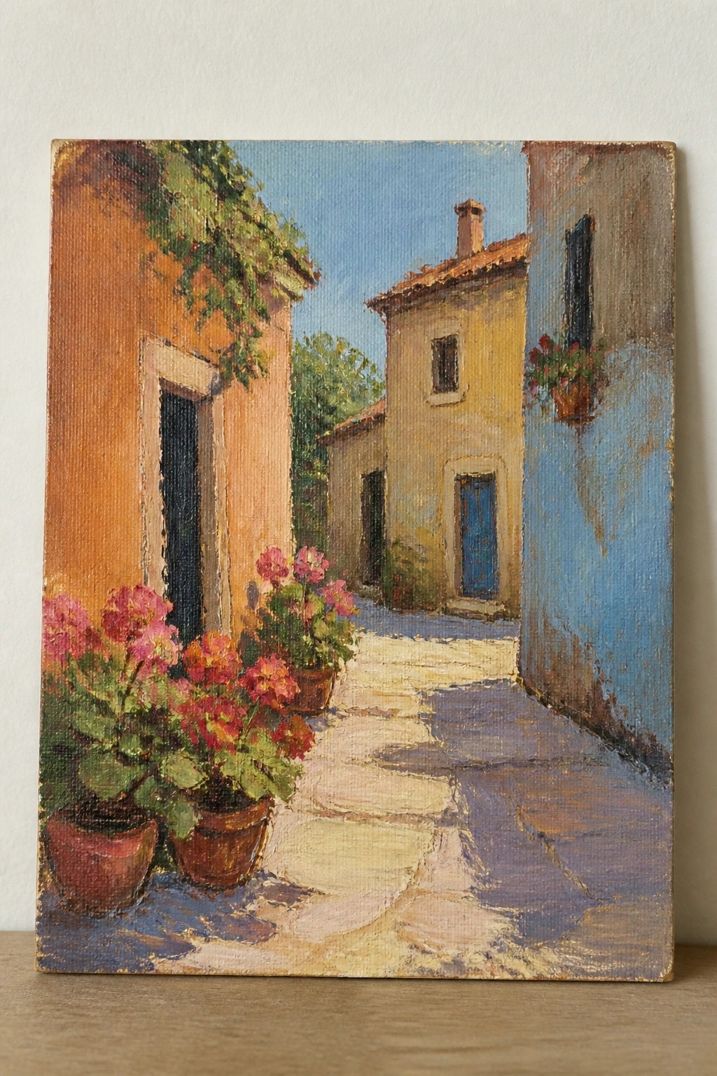 Narrow sunlit alley with potted pink flowers beside peach, yellow, and blue buildings