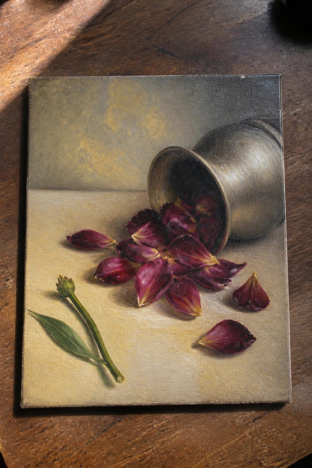 Still life of overturned brass cup spilling dark purple petals beside a green stem.