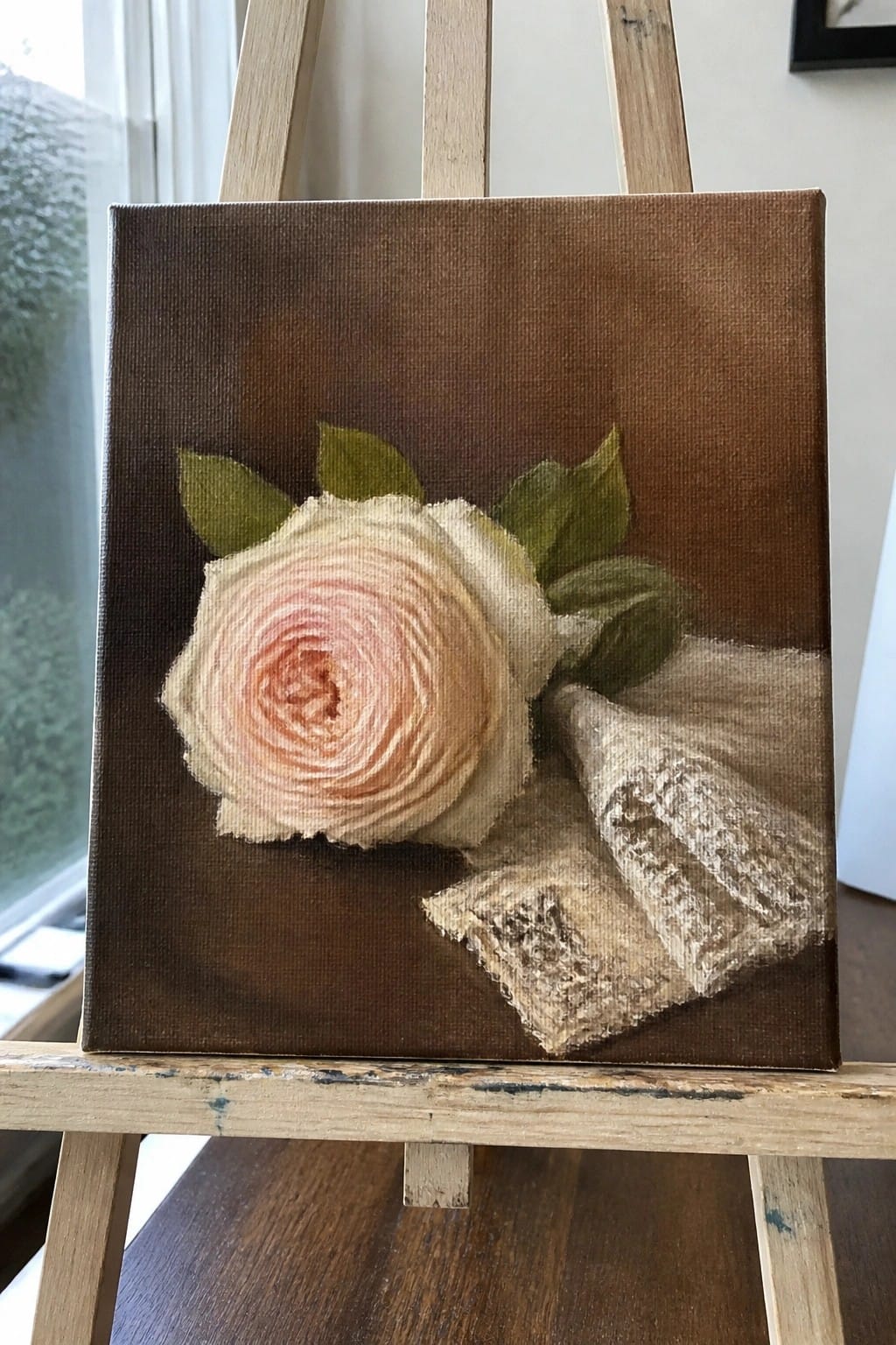 Pale pink rose painting with leaves and lace fabric on wooden easel.