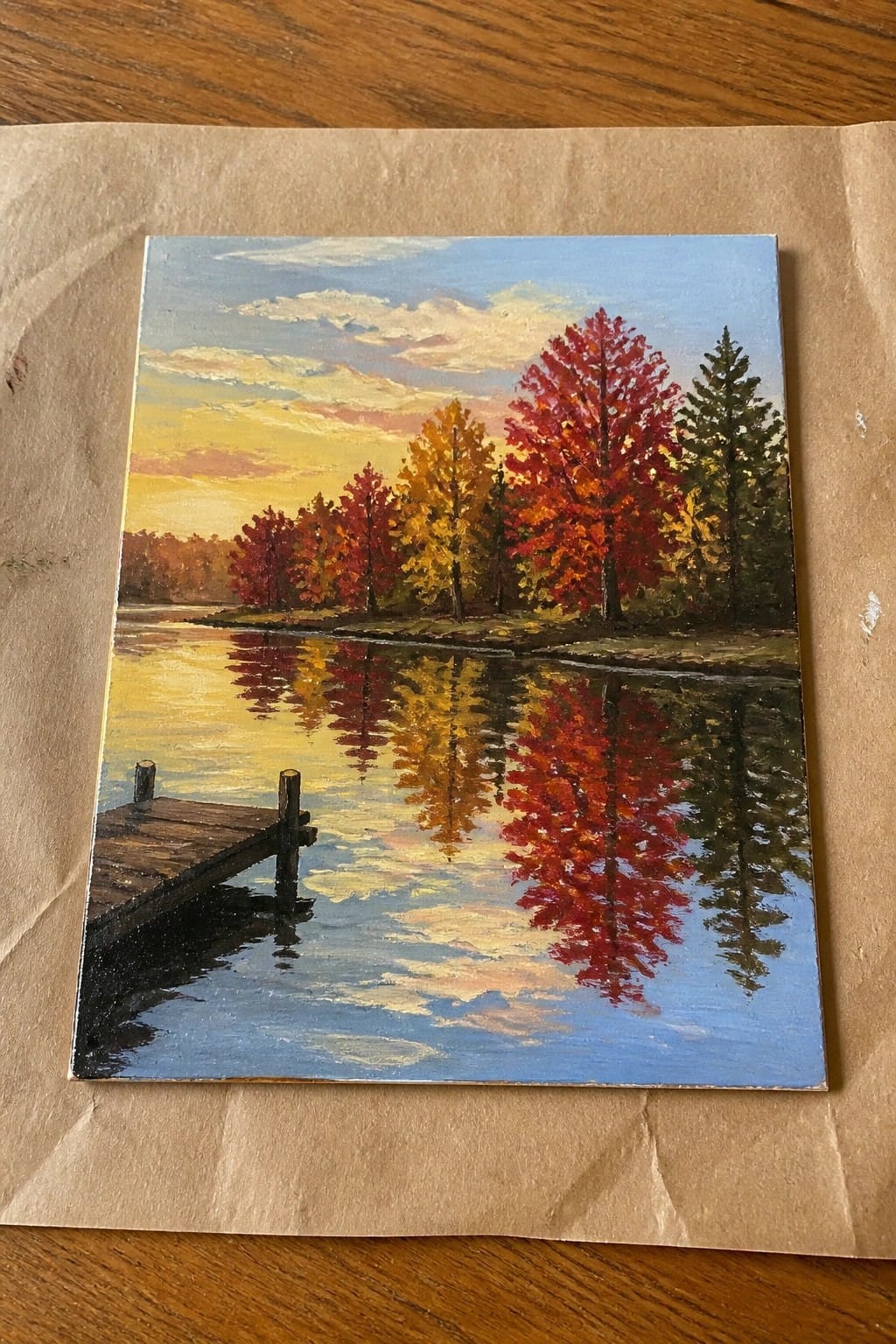 Colorful autumn painting with red-orange trees reflected in calm lake beside wooden dock.