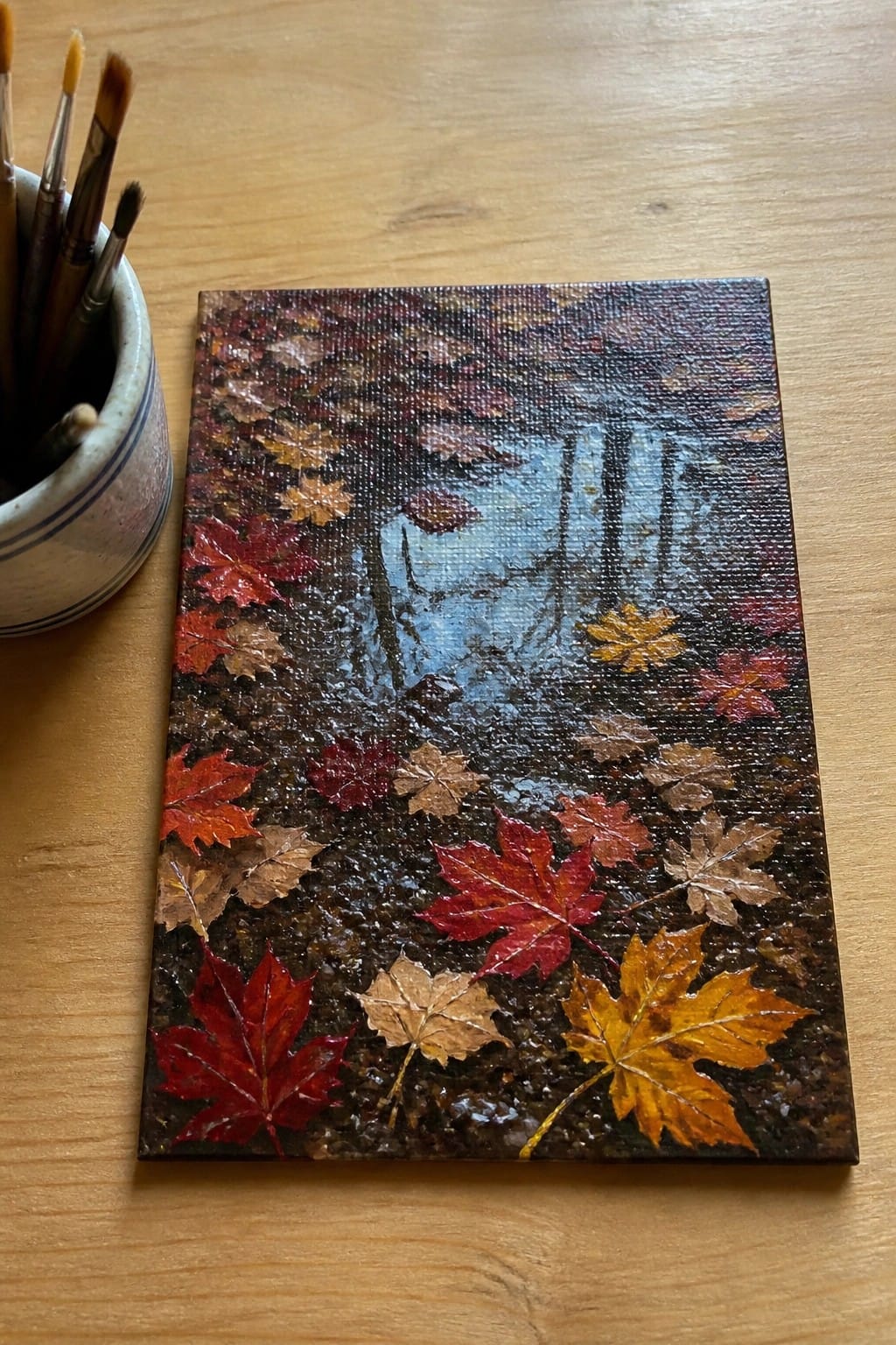 Small canvas autumn forest painting with leaves beside paintbrushes on wooden table