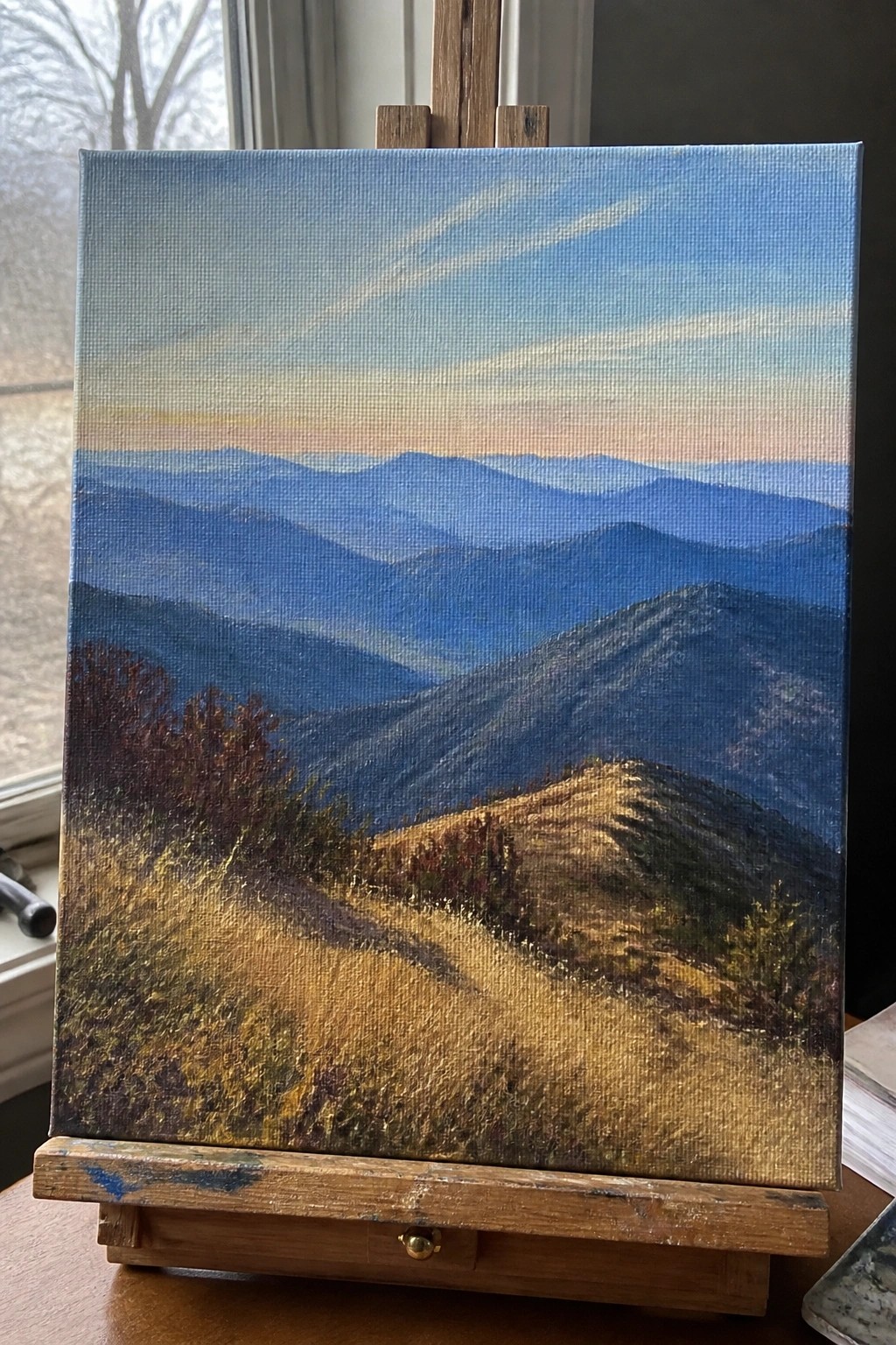 Oil painting of layered blue mountains and golden hills on wooden easel by window