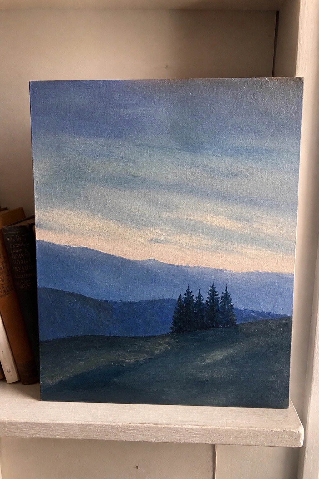 Blue landscape painting with layered hills and pine trees on white shelf beside books