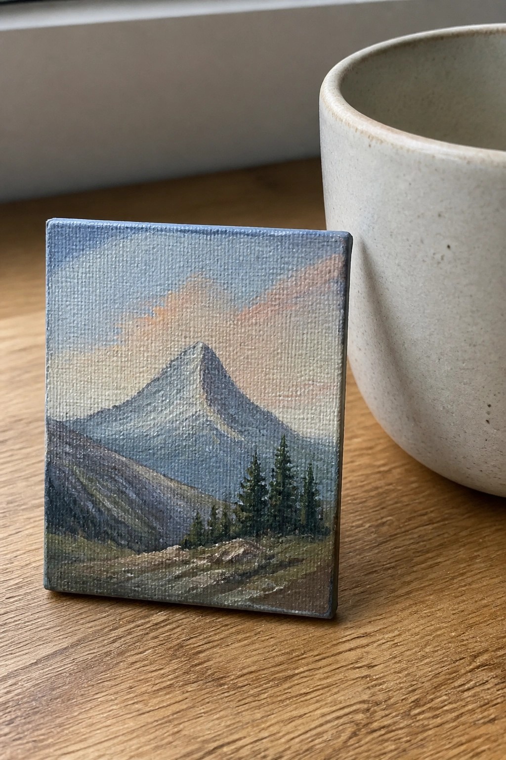 Small oil painting of a mountain peak with pine trees and a path under a blue sky with orange clouds, on a wooden table next to a beige cup.