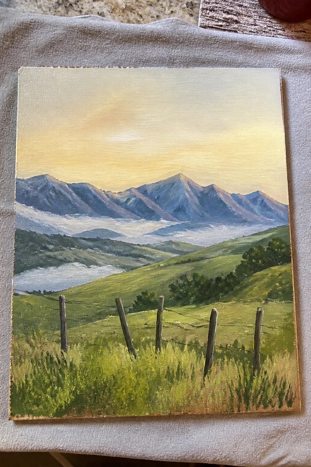 Painted landscape of green hills with wooden fence overlooking misty mountains under golden sky