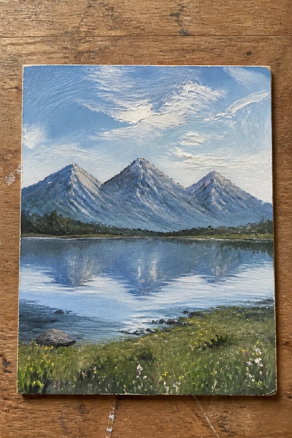 Painting of jagged mountains reflected in a calm blue lake with wildflower meadow.