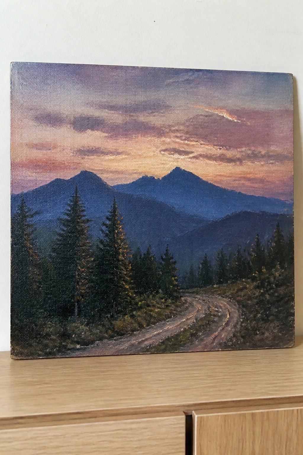 Oil painting of winding dirt road through pines toward mountains under colorful sunset sky.