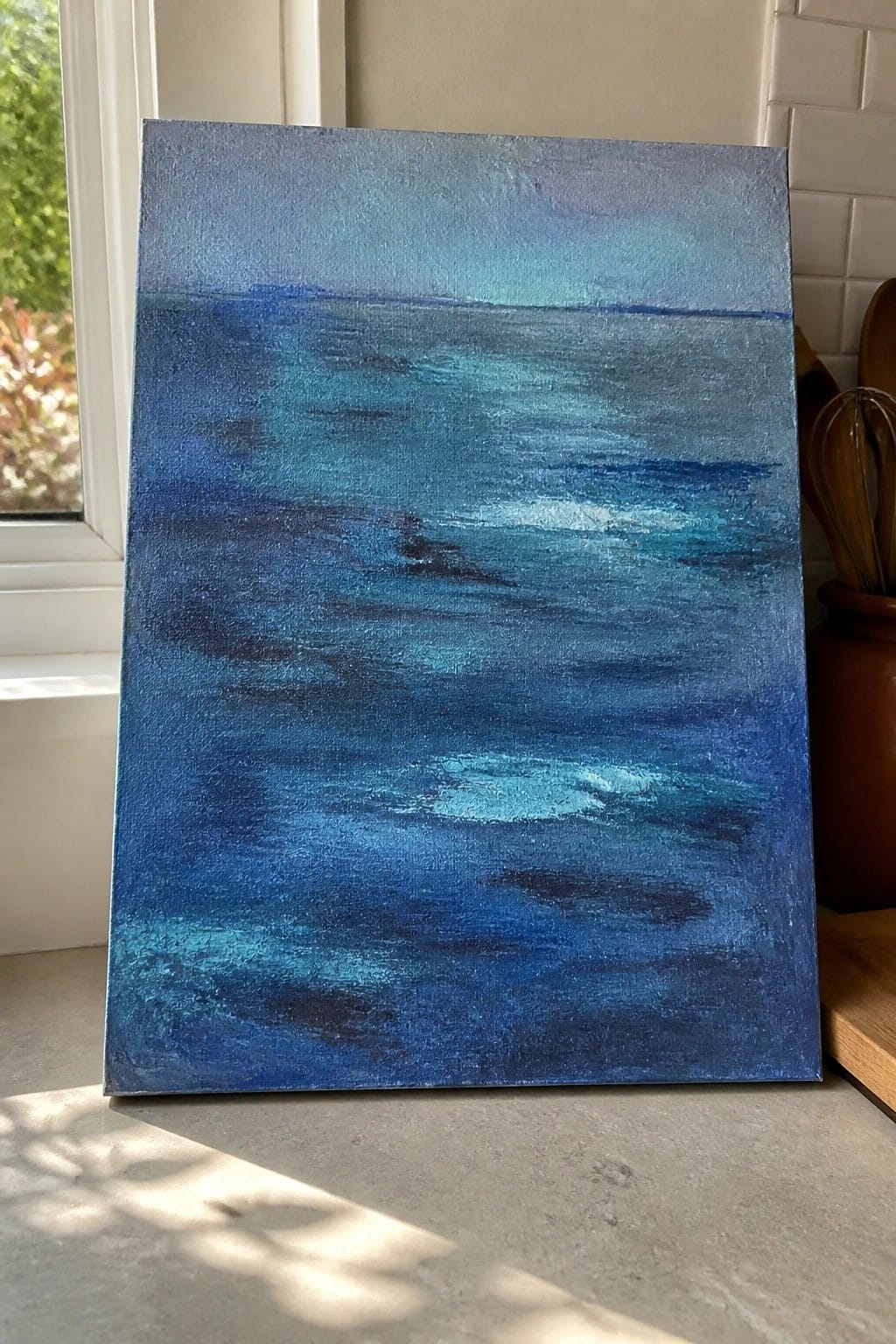 Abstract blue ocean painting with waves leaning against kitchen wall near window.