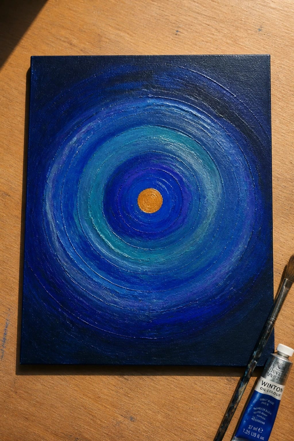 Concentric blue circles with gold center on canvas beside paint tube and brush on wood.