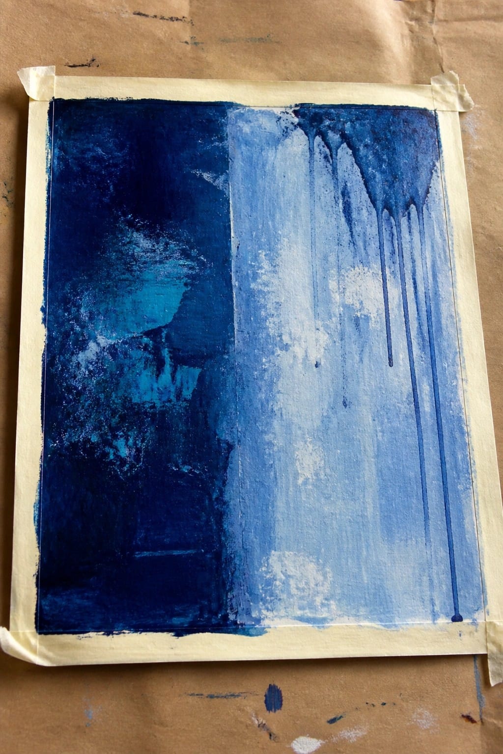 Abstract blue painting taped to brown surface, dark textures left with vertical drips right.