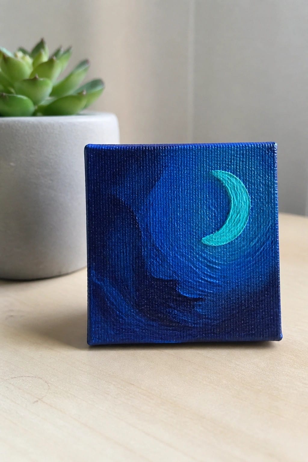 Small canvas painting of turquoise crescent moon on swirling blue night sky beside succulent.