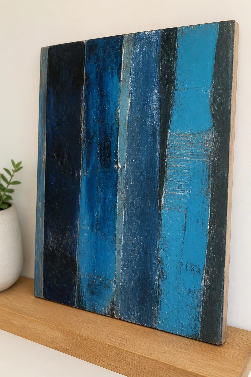 A square abstract oil painting with vertical stripes in multiple shades of blue on a wooden shelf.