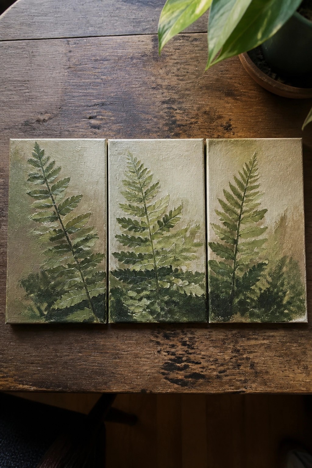 Three canvas paintings of ferns in greens on wooden table.