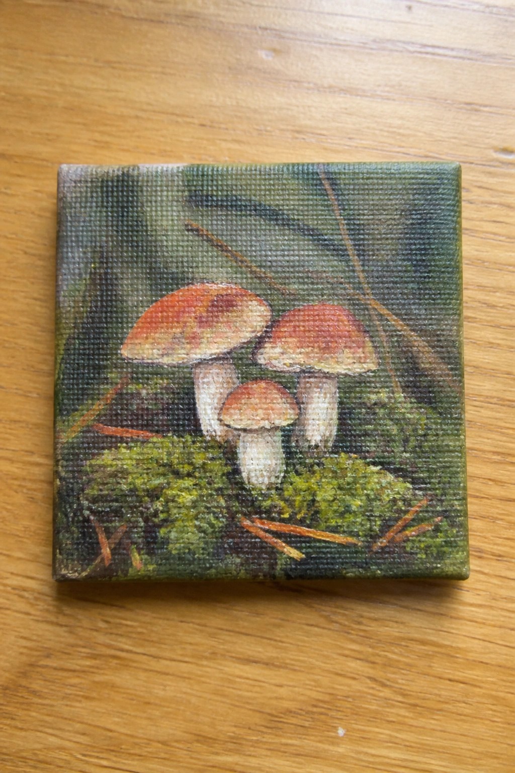 Three reddish-capped mushrooms painted on small canvas atop wooden surface.
