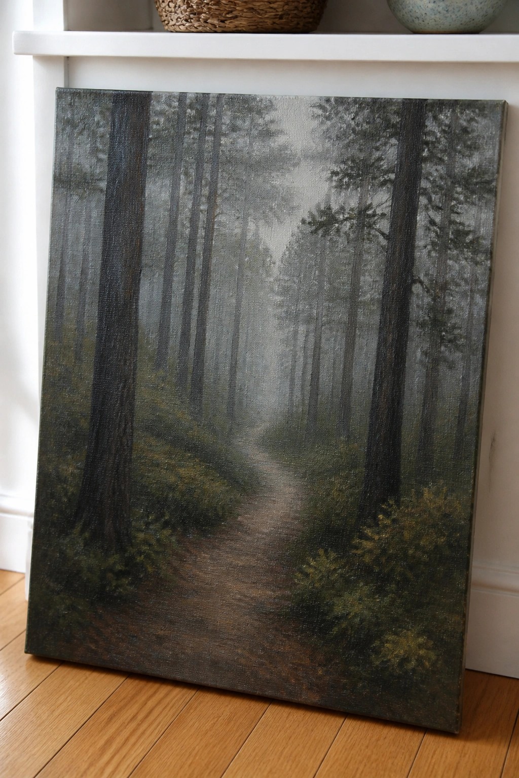 Misty forest path painting on canvas leaning against white shelf on wood floor.