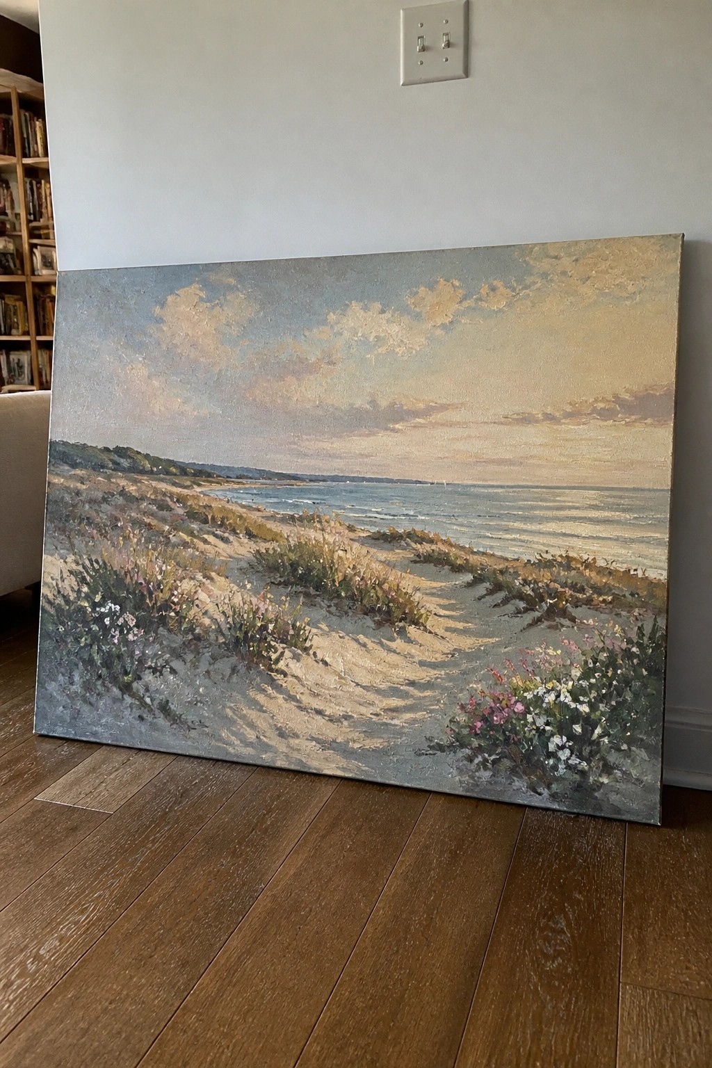 Large beach landscape painting with dunes and wildflowers leaning on wooden floor against wall