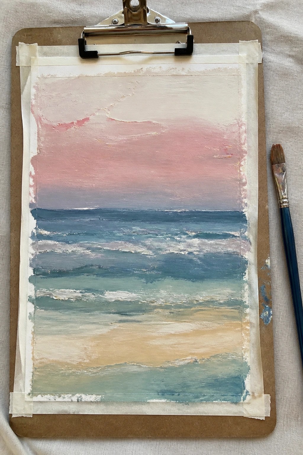 Seascape painting on clipboard with pink sky, blue waves, sandy shore and paintbrush nearby