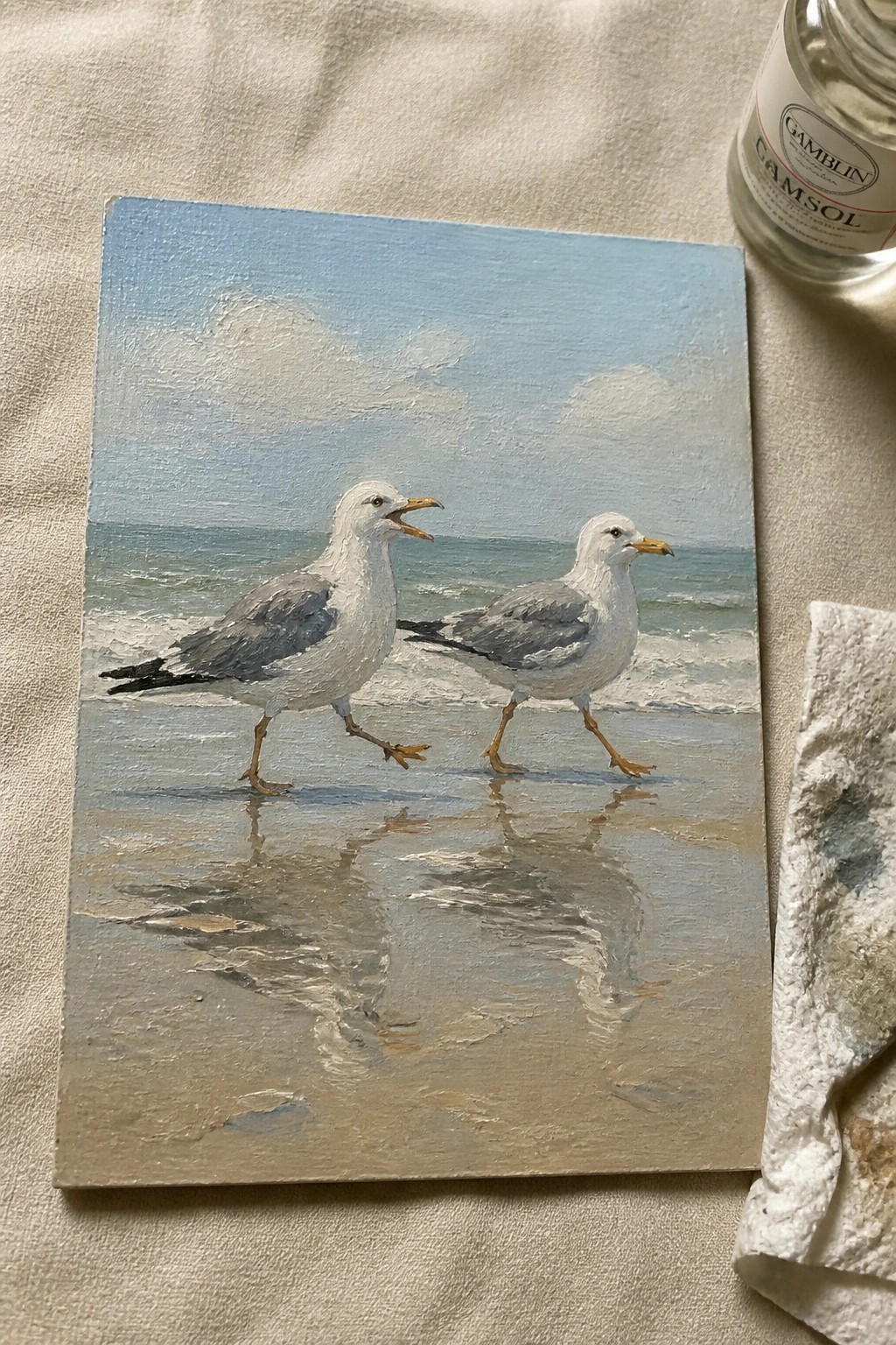 Two seagulls on wet beach with ocean reflections, beside Gamsol bottle and rag