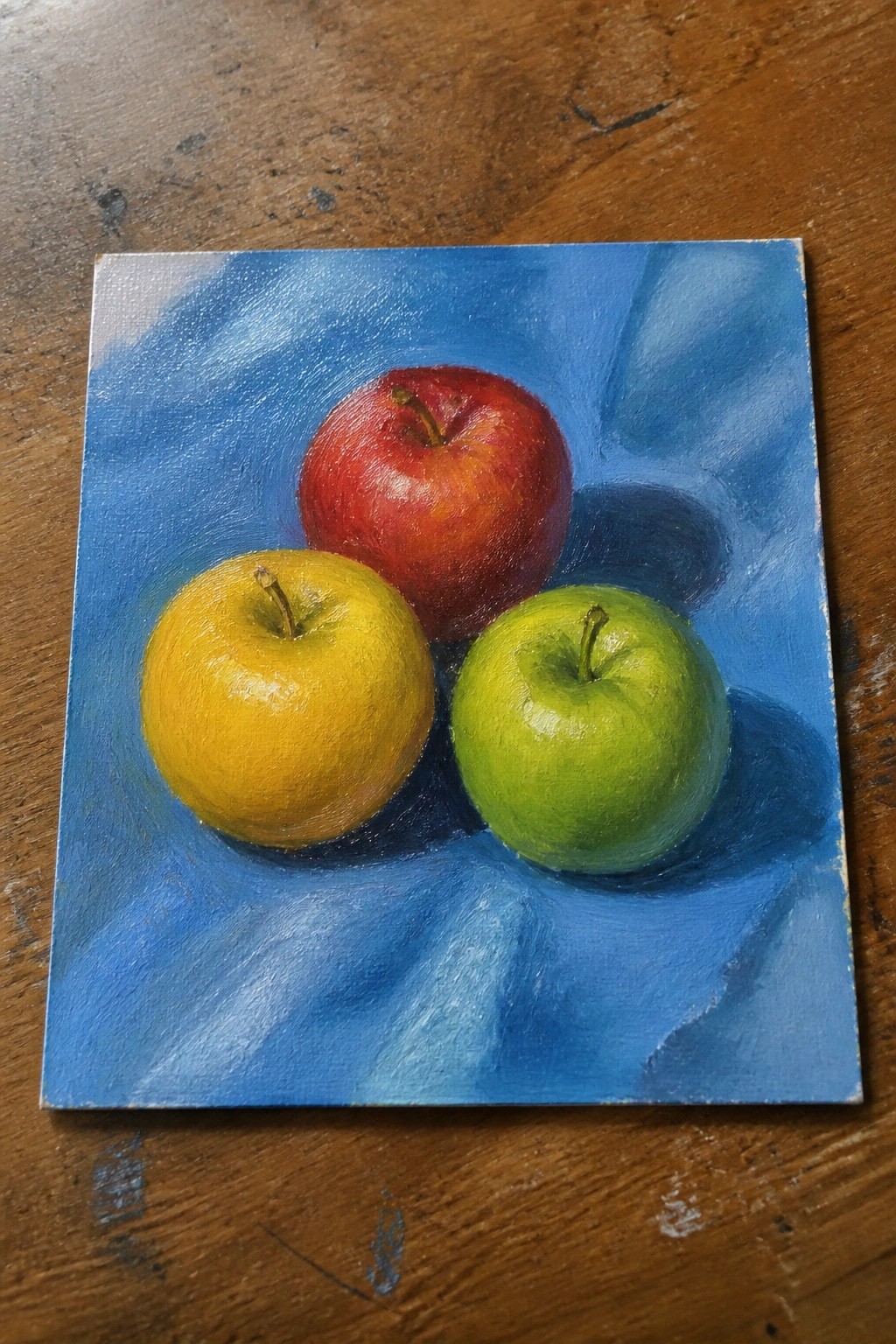 Three apples—red, yellow, and green—on draped blue fabric in oil painting.