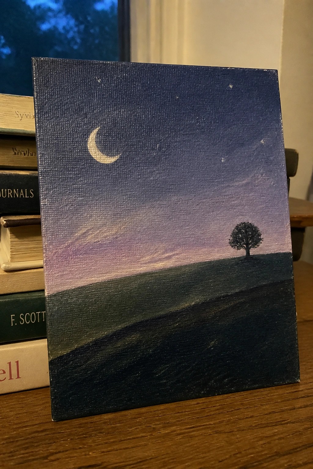 Acrylic painting of crescent moon over purple starry sky with lone tree on hill against books.