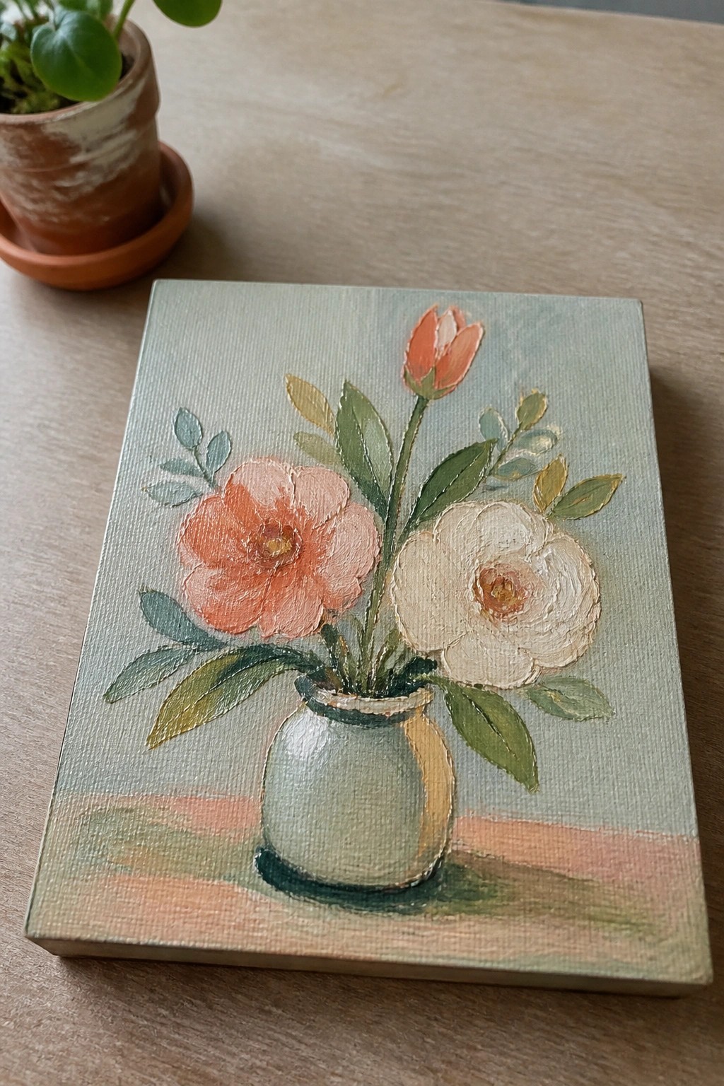 Small oil painting of coral, cream, and orange flowers in a pale vase on a wooden table.