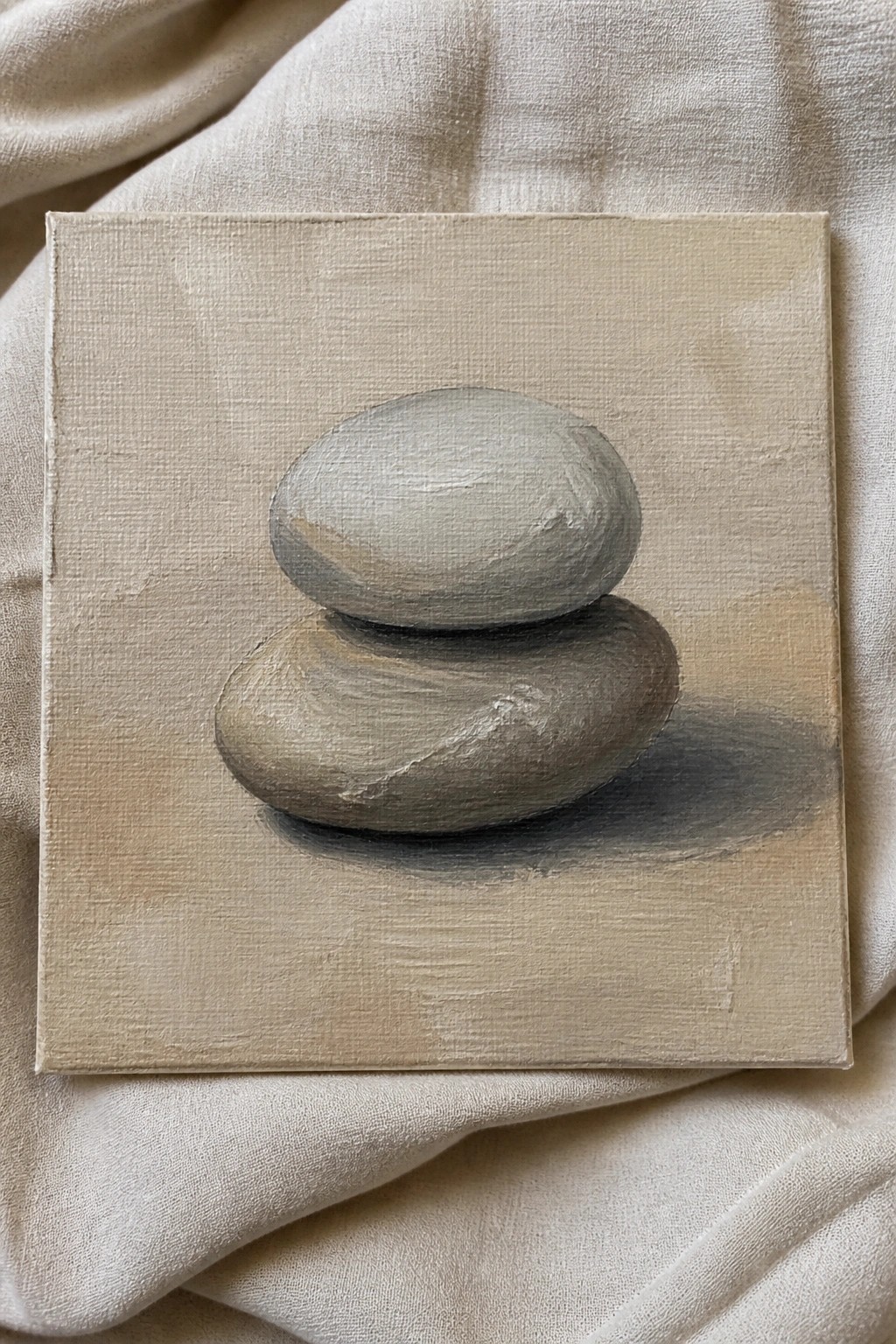 Painting of two stacked gray stones on beige canvas over wrinkled fabric.