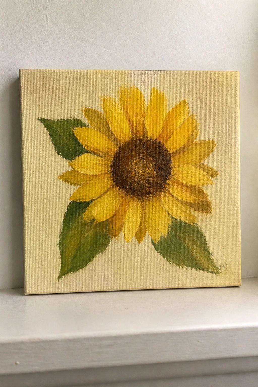 Square canvas painting of yellow sunflower with green leaves on white shelf