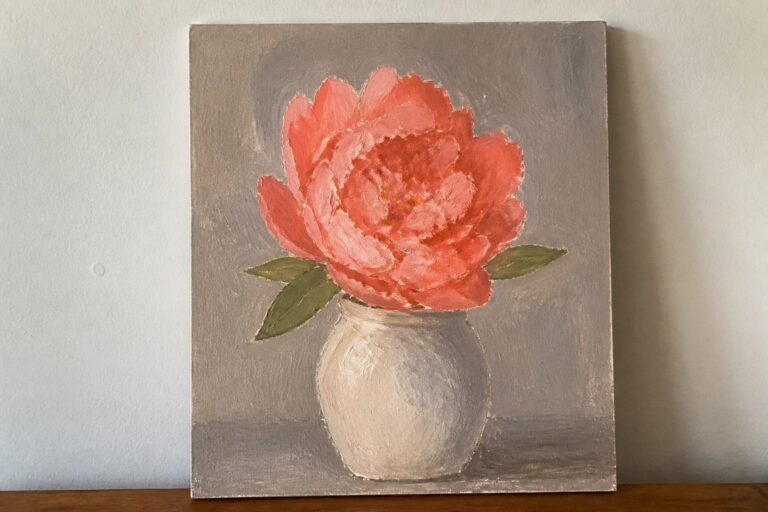Pink peony in a white vase on wooden surface against muted gray background.