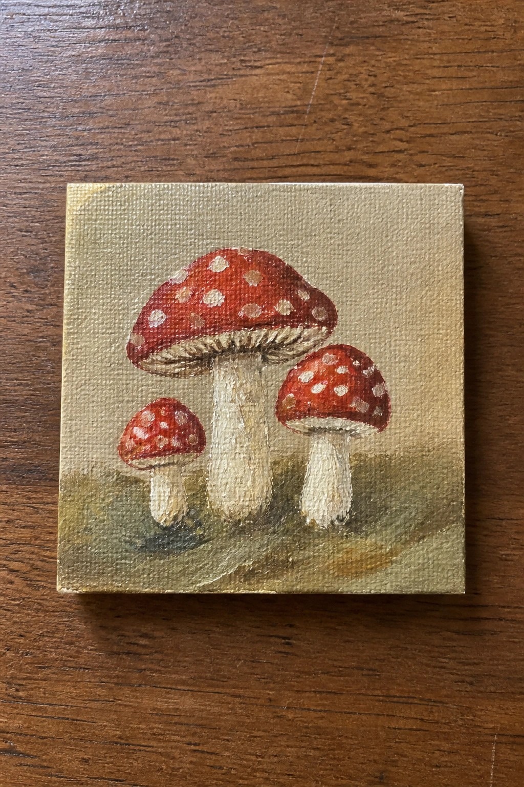 Three red mushrooms with white spots painted on small beige canvas