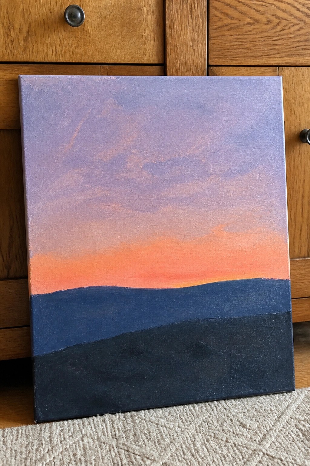 Abstract sunset painting with purple-orange sky over dark blue and black hills on canvas against wood