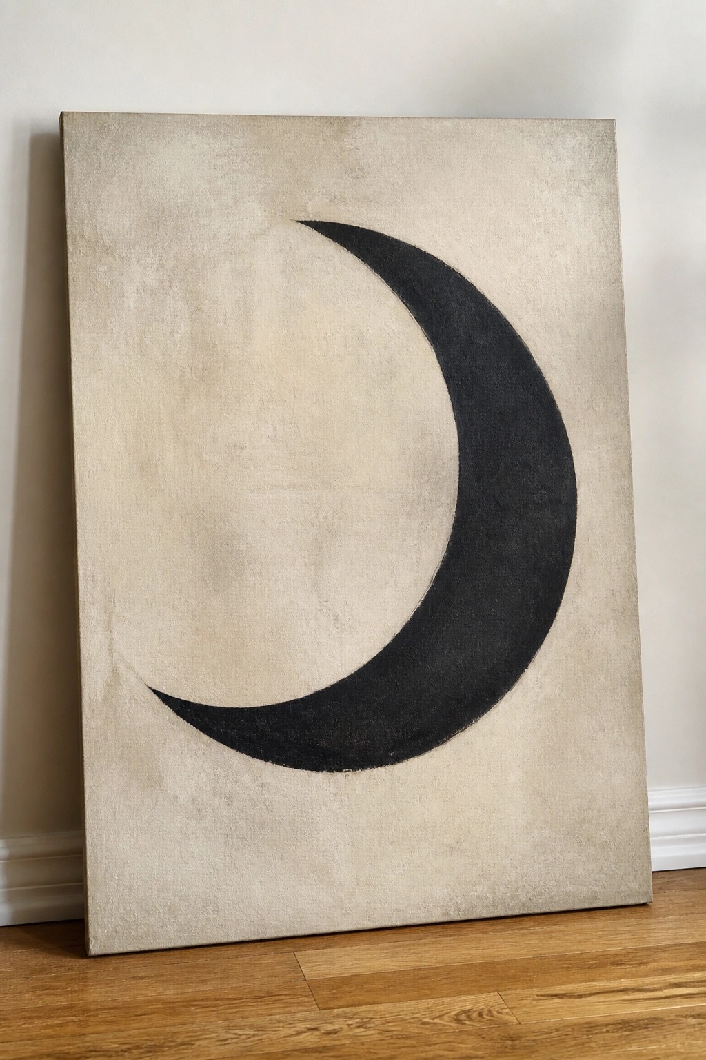 Black crescent moon on textured beige canvas propped against white wall on wood floor.