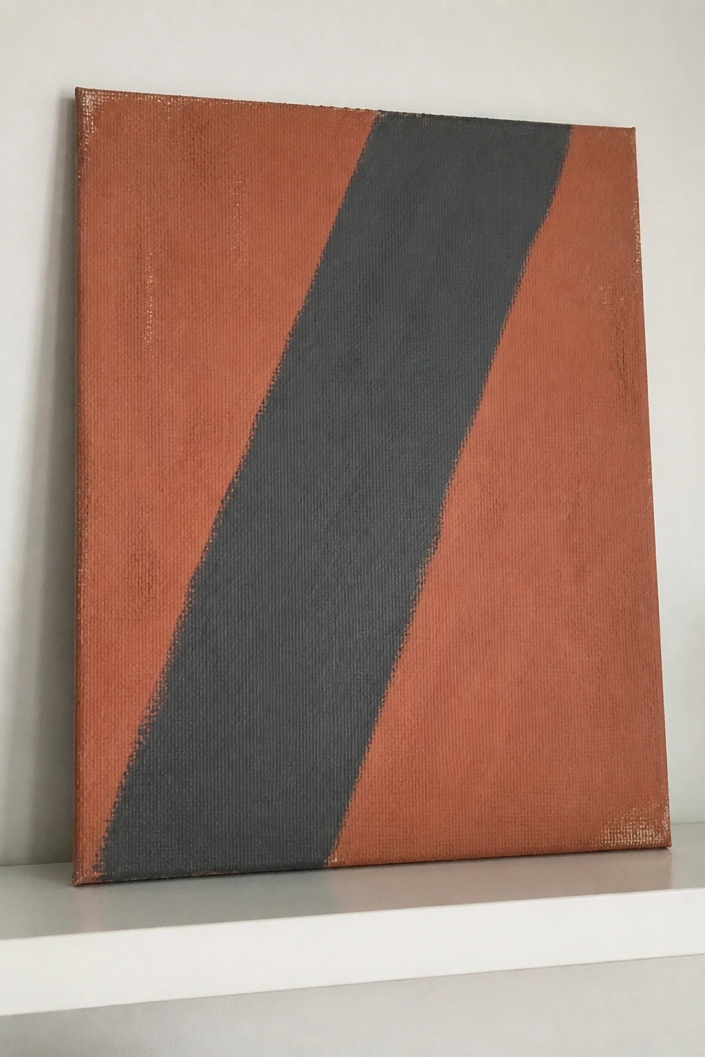 Abstract canvas with dark gray diagonal stripe on rust-orange background on white shelf