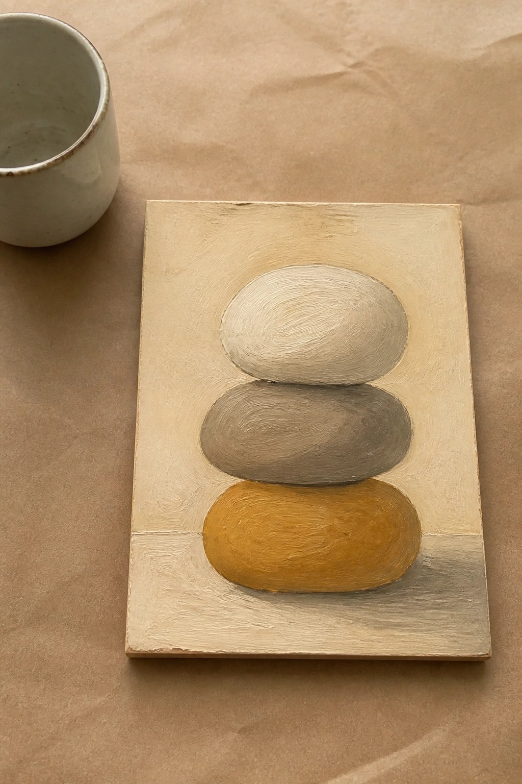Stacked white, gray, and ochre stones painted on small beige canvas beside ceramic cup.