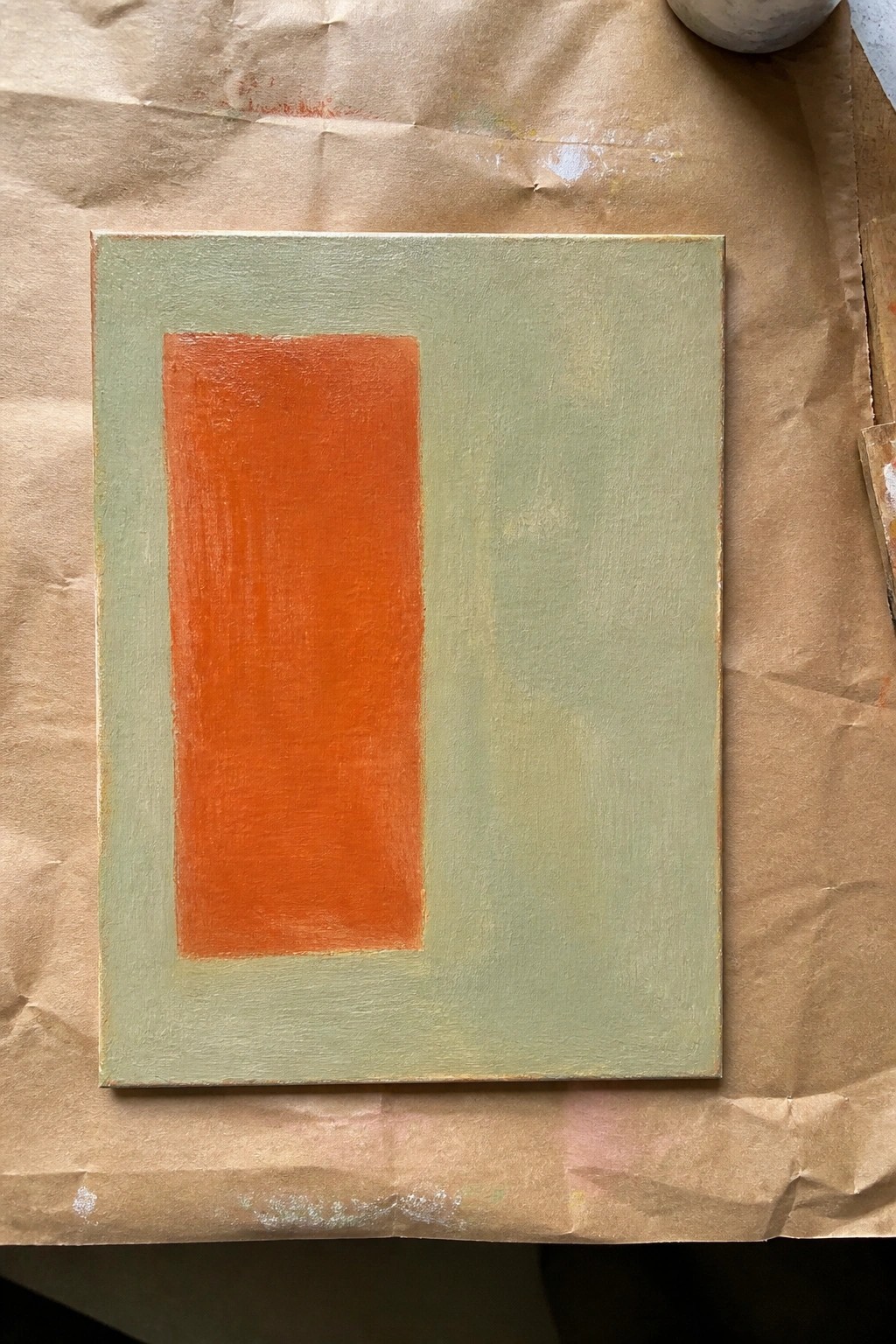 Rectangular canvas with orange block on green background placed on stained brown paper.
