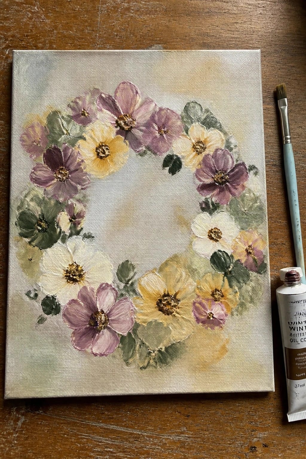 Floral wreath painting on canvas beside blue brush and oil paint tube on wood