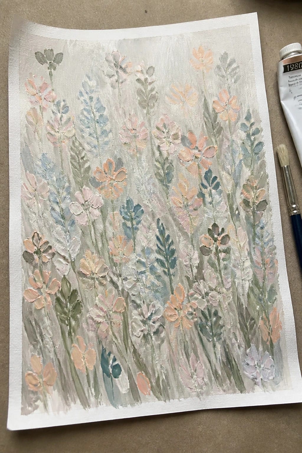 Watercolor of soft pastel wildflowers beside paint tube and brush on paper