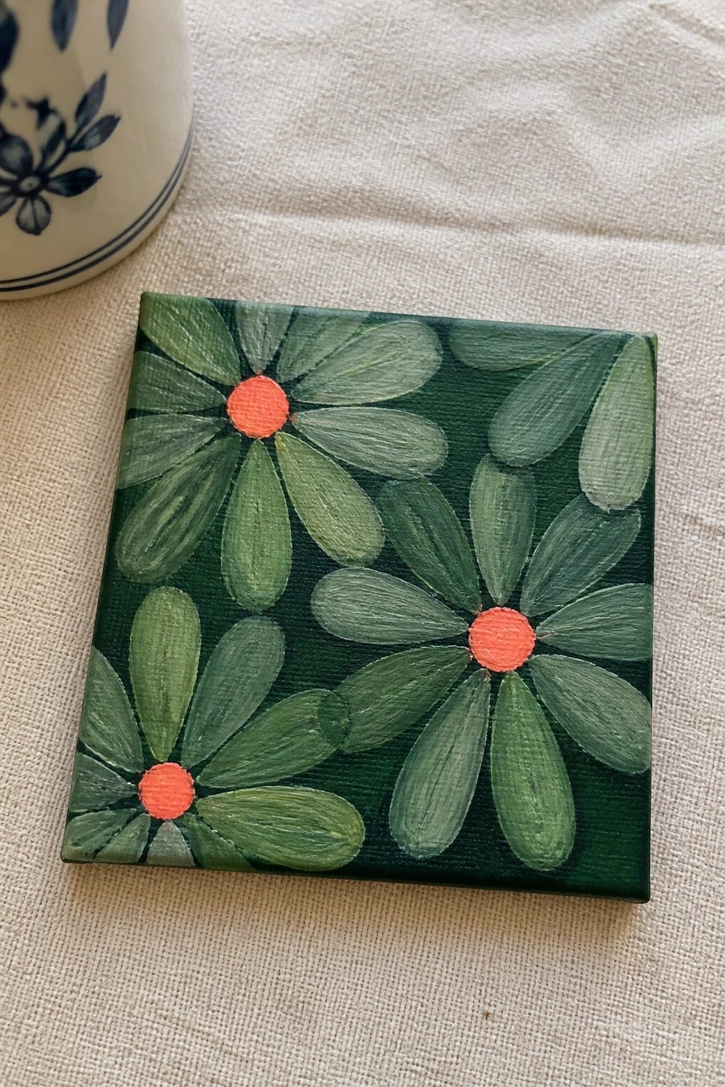 Three green-petaled flowers with orange centers on small square canvas beside ceramic mug