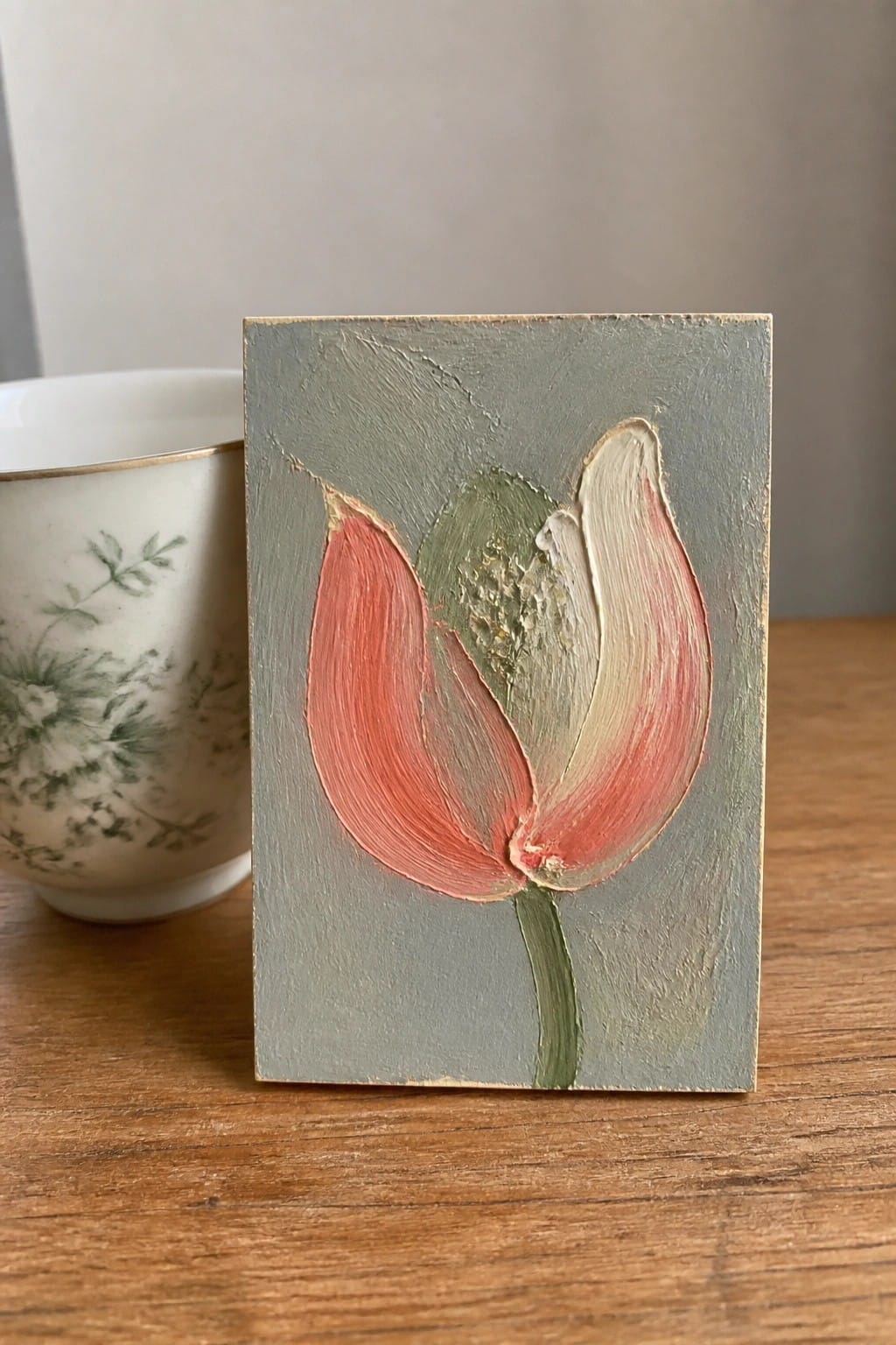 A small oil painting of a coral tulip with visible brushstrokes on a gray background sits on a wooden table beside a white floral teacup.