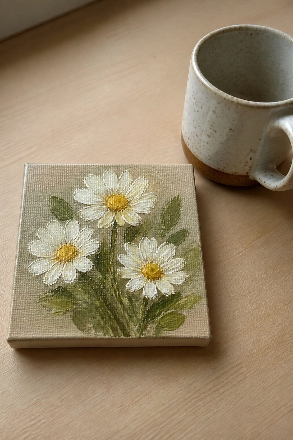 Small canvas with three white daisies beside a speckled ceramic mug on wood.