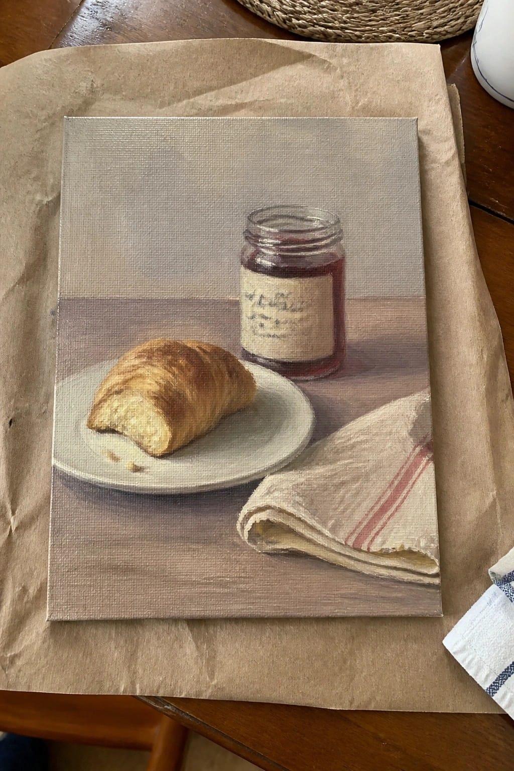 A realistic oil painting of a croissant on a white plate next to a glass jam jar and a folded cloth napkin on a wooden table.