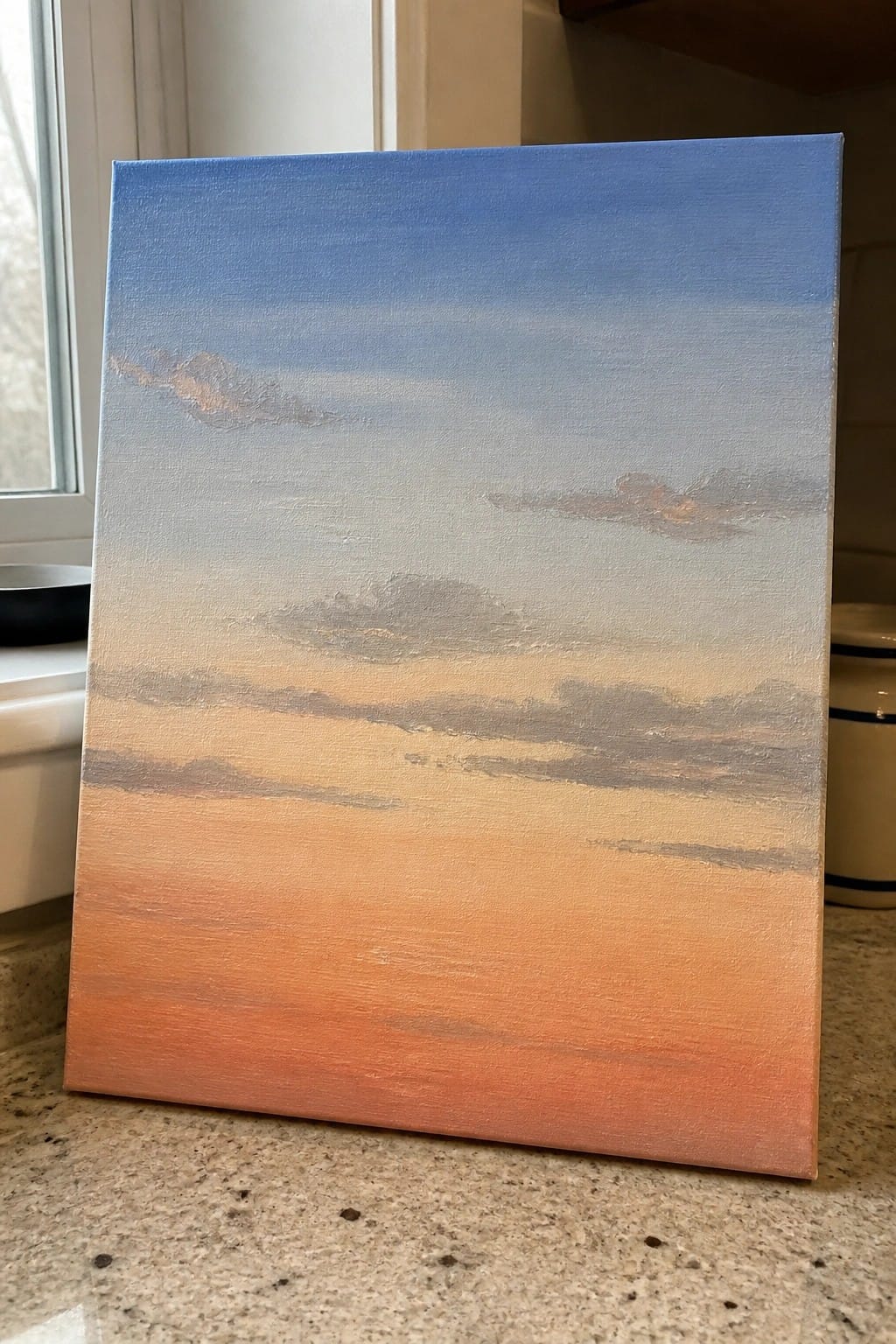 Canvas painting of blue-orange gradient sky with layered gray clouds on countertop.