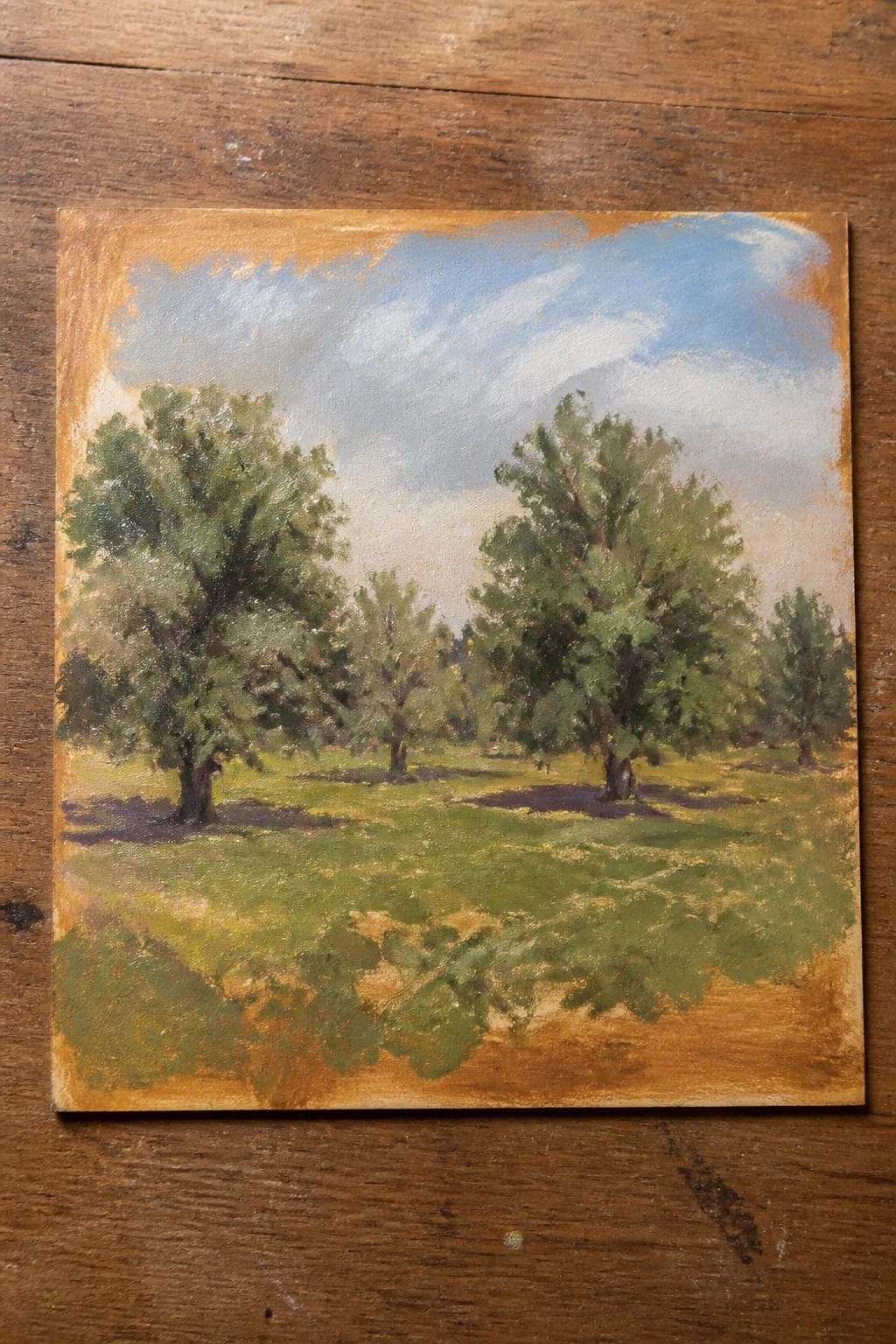 An oil painting of several green trees in a grassy field under a blue sky, shown on a wooden surface.