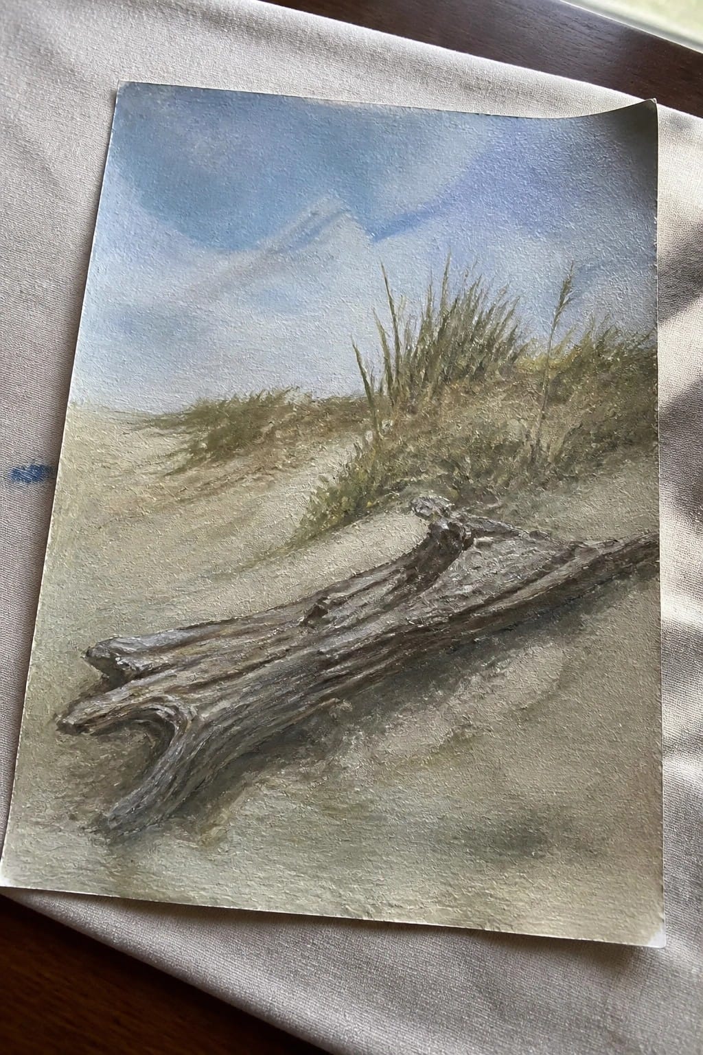 Driftwood log on sandy dunes with grasses under blue cloudy sky in landscape painting