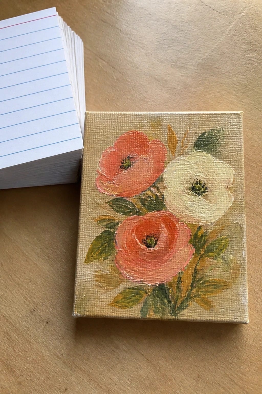 Small canvas painting of coral and white flowers beside stack of lined cards on wood.