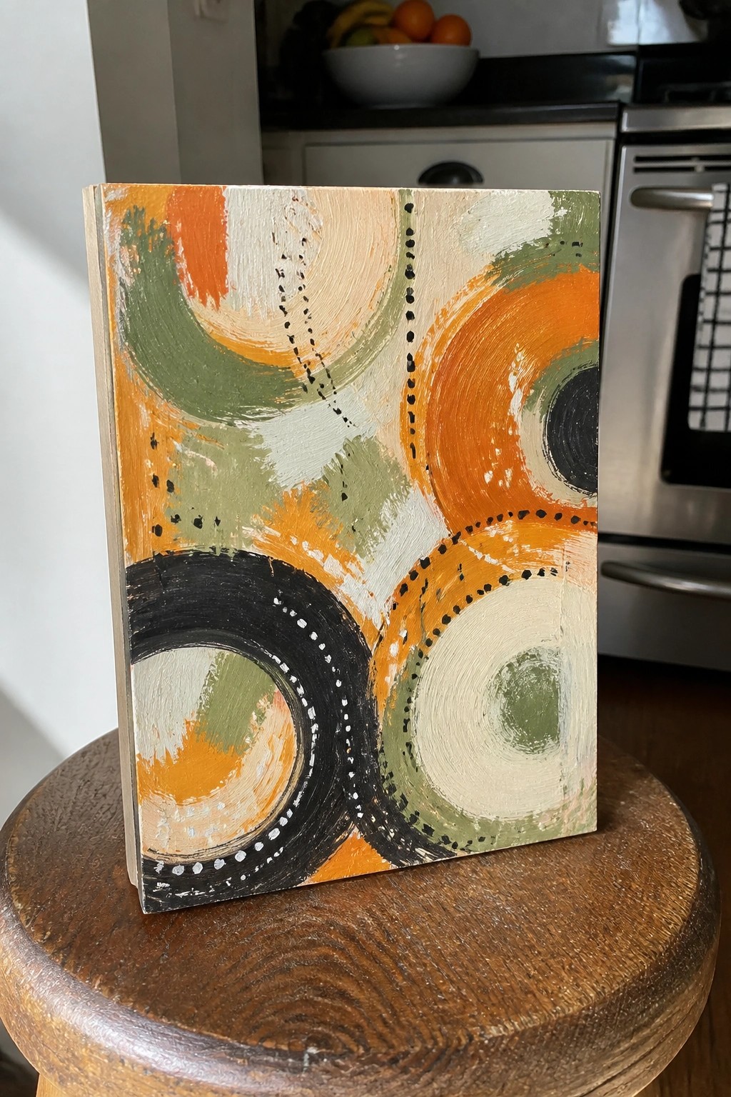 Abstract painting with orange, green, and black circles resting on a wooden stool.