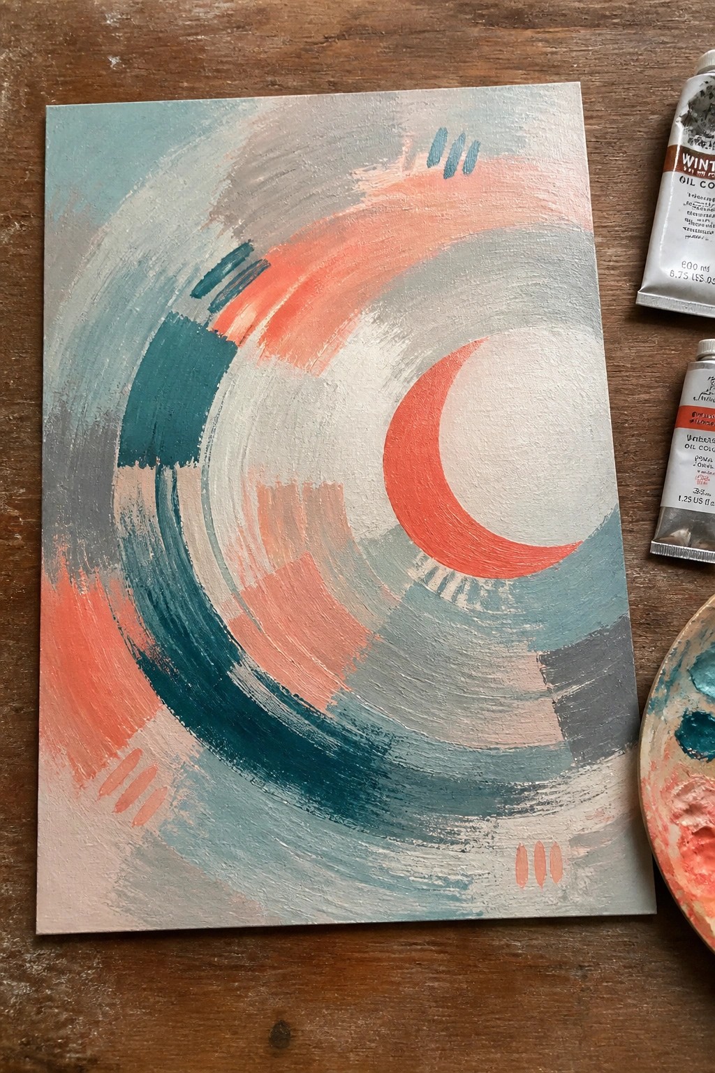 Abstract canvas with teal coral gray swirls, red crescent, white circle, and paint tubes on wood